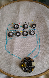 Fabric in a hoop, with a microcontroller and sevel LEDs pinned in place.  Traces are drawn in fabric marker to show where conductive thread will be stitched.